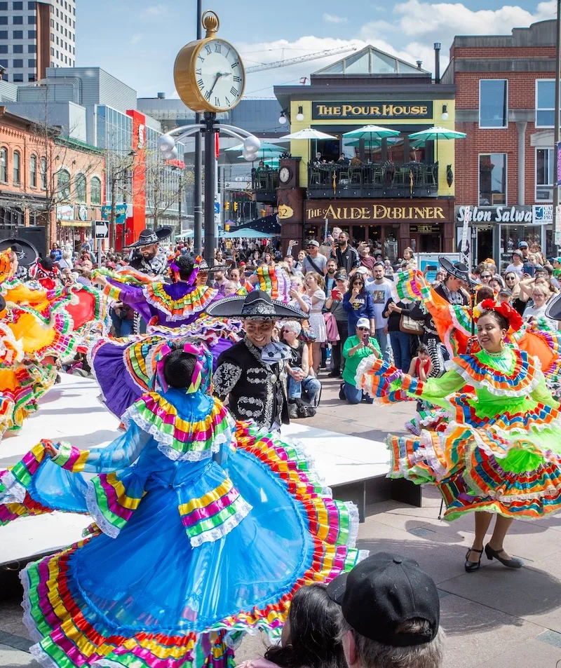 Dancers in colorful traditional Mexican dresses perform outdoors for a crowd in a lively city square, with vibrant buildings, a clock, and restaurants in the background.