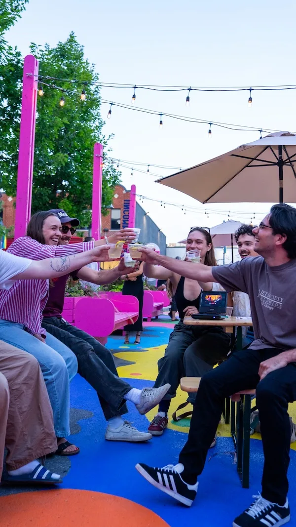 A group of friends sitting at a colorful outdoor area, smiling and raising their drinks together for a toast under string lights, with pink benches and umbrellas in the background.