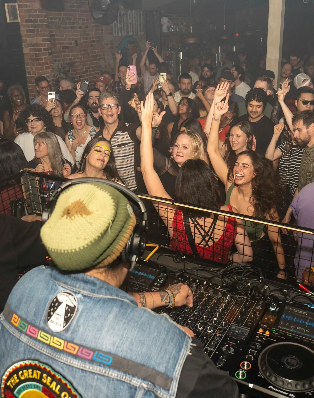 A DJ wearing a beanie plays music for a lively crowd dancing and smiling in a dimly lit club. Many people have their hands up, enjoying the energetic atmosphere.