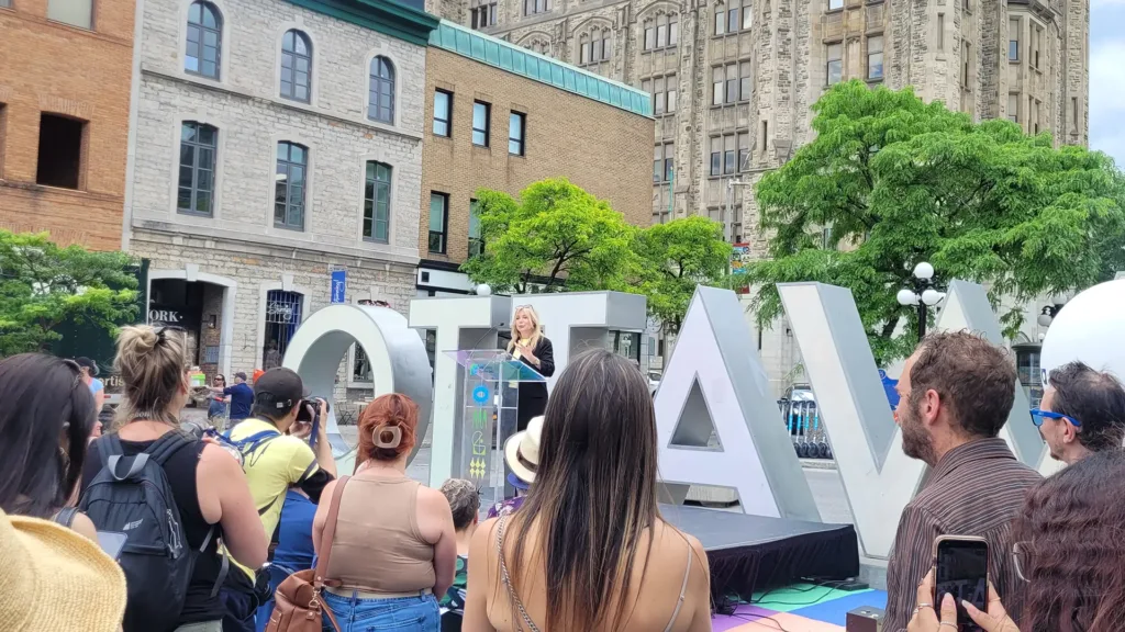 A diverse crowd gathers around a large sign displaying the word "Ottawa" in bold letters