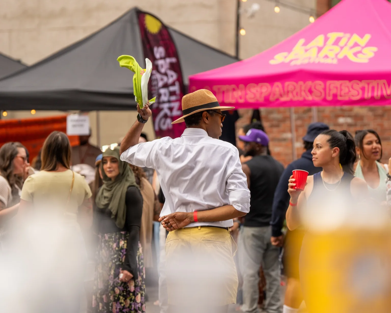 A man in a hat and white shirt dances with a green scarf at an outdoor festival. People stand around him, some holding drinks, with colorful tents and a Latin Sparks Festival sign in the background.